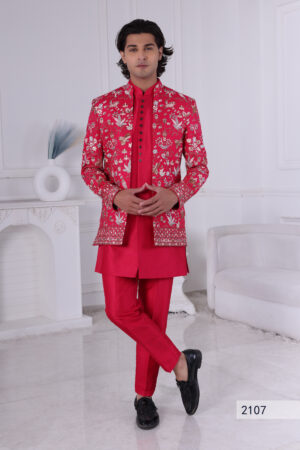 Man wearing a red embroidered Jodhpuri suit with floral patterns, gold and silver embroidery, paired with a red kurta. The background features a minimalist white interior with soft lighting.