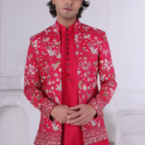 Man wearing a red embroidered Jodhpuri suit with floral patterns, gold and silver embroidery, paired with a red kurta. The background features a minimalist white interior with soft lighting.