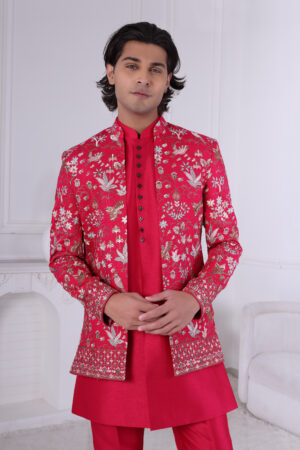 Man wearing a red embroidered Jodhpuri suit with floral patterns, gold and silver embroidery, paired with a red kurta. The background features a minimalist white interior with soft lighting.