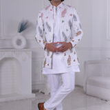 Man wearing a white Jodhpuri suit with multicolor leaf and butterfly prints, standing indoors with folded arms.
