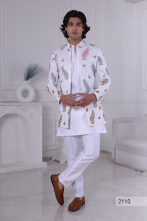 Man wearing a white Jodhpuri suit with multicolor leaf and butterfly prints, standing indoors with folded arms.