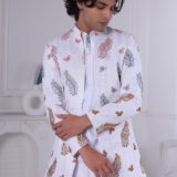 Man wearing a white Jodhpuri suit with multicolor leaf and butterfly prints, standing indoors with folded arms.
