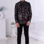 Man wearing a black embroidered Jodhpuri suit with multicolor floral and bird motifs, posing indoors with hands clasped.