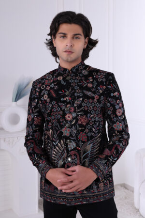 Man wearing a black embroidered Jodhpuri suit with multicolor floral and bird motifs, posing indoors with hands clasped.