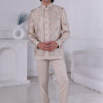 Man wearing a beige Jodhpuri suit with pastel floral embroidery and metallic buttons, posing indoors.