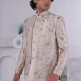 Man wearing a beige Jodhpuri suit with pastel floral embroidery and metallic buttons, posing indoors.