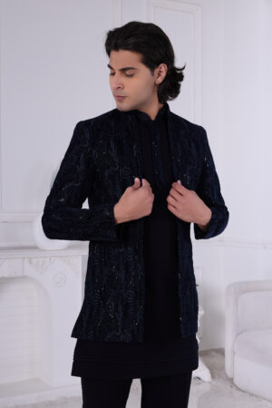 Man wearing a navy blue textured Jodhpuri suit with subtle embroidery and sequins, posing indoors with hands on jacket.
