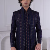Man wearing a navy blue Jodhpuri suit with embroidered vertical stripes and mirror accents, standing indoors.
