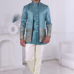 Man wearing a teal blue embroidered Jodhpuri suit with golden embroidery, mirror accents, and traditional motifs, posing indoors.
