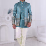 Man wearing a teal blue embroidered Jodhpuri suit with golden embroidery, mirror accents, and traditional motifs, posing indoors.
