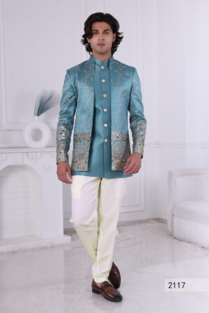 Man wearing a teal blue embroidered Jodhpuri suit with golden embroidery, mirror accents, and traditional motifs, posing indoors.