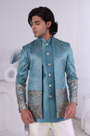 Man wearing a teal blue embroidered Jodhpuri suit with golden embroidery, mirror accents, and traditional motifs, posing indoors.