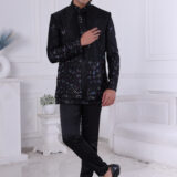 Man wearing a black embroidered Jodhpuri suit with multicolor floral patterns and studded detailing, posing indoors.