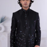 Man wearing a black embroidered Jodhpuri suit with multicolor floral patterns and studded detailing, posing indoors.
