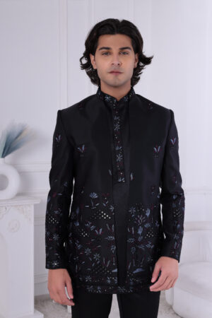 Man wearing a black embroidered Jodhpuri suit with multicolor floral patterns and studded detailing, posing indoors.