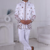 Man wearing a white embroidered Jodhpuri suit with floral, bird, and deer motifs, standing indoors.
