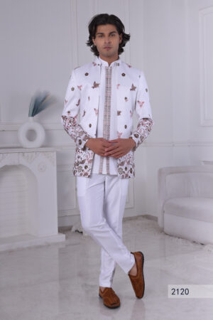 Man wearing a white embroidered Jodhpuri suit with floral, bird, and deer motifs, standing indoors.