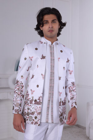Man wearing a white embroidered Jodhpuri suit with floral, bird, and deer motifs, standing indoors.