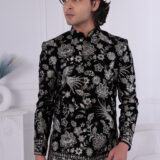 Man wearing a black velvet embroidered Jodhpuri suit with metallic floral patterns, posing indoors.