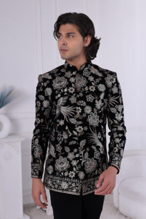 Man wearing a black velvet embroidered Jodhpuri suit with metallic floral patterns, posing indoors.