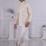 Man wearing a cream Jodhpuri suit with textured patterns, posing indoors with hands clasped.