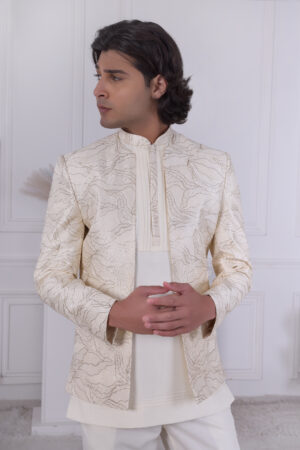 Man wearing a cream Jodhpuri suit with textured patterns, posing indoors with hands clasped.