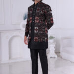 Man wearing a black Jodhpuri suit with maroon and grey patterned embroidery, looking down with one hand on chest.