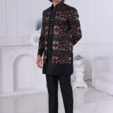 Man wearing a black Jodhpuri suit with maroon and grey patterned embroidery, looking down with one hand on chest.