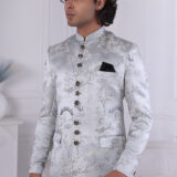 Man wearing a silver brocade Jodhpuri suit with intricate patterns, ornate buttons, and a black pocket square.