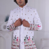 Man wearing a white Jodhpuri suit featuring golden embroidery and colorful deer motifs on the cuffs and hemline.