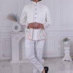 A man wearing a white Jodhpuri suit with silver buttons, sparkling texture, and pastel flamingo embroidery at the hem.