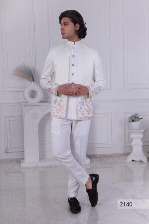 A man wearing a white Jodhpuri suit with silver buttons, sparkling texture, and pastel flamingo embroidery at the hem.