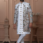 Men’s white Indo-Western outfit with embroidered jacket in floral and peacock designs, paired with plain kurta and churidar.