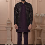 A man wearing a maroon Indo-Western outfit with an intricately embroidered mosaic-patterned jacket, standing against a peach-toned classic interior backdrop.