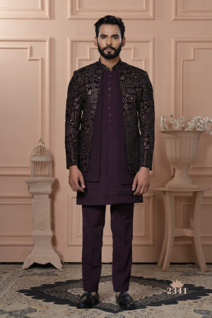 A man wearing a maroon Indo-Western outfit with an intricately embroidered mosaic-patterned jacket, standing against a peach-toned classic interior backdrop.