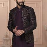 Man wearing a maroon Indo-Western outfit with an intricately embroidered mosaic-patterned jacket, standing against a peach-toned classic interior backdrop.