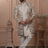 Man dressed in an ivory Indo-Western outfit with a floral and palm leaf embroidered jacket, standing in front of a peach-toned, classic interior backdrop.