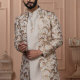 Man dressed in an ivory Indo-Western outfit with a floral and palm leaf embroidered jacket, standing in front of a peach-toned, classic interior backdrop.