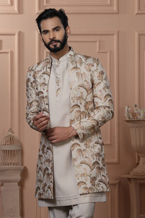 Man dressed in an ivory Indo-Western outfit with a floral and palm leaf embroidered jacket, standing in front of a peach-toned, classic interior backdrop.