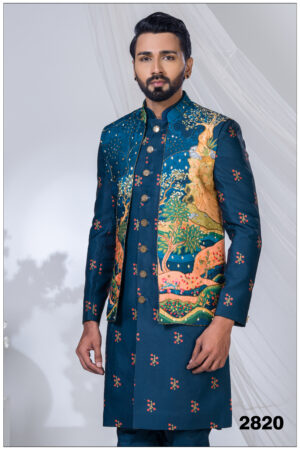 A man wearing a royal blue open Indo-Western suit with artistic landscape embroidery in gold, green, and coral tones, paired with metallic buttons.