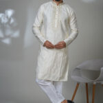 Man wearing an ivory embroidered kurta with intricate front placket and all-over floral accents, styled with white pants and black formal shoes.