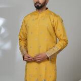 Man wearing a mustard yellow embroidered kurta with silver floral detailing, paired with white pants and black shoes, standing in a modern studio setup.