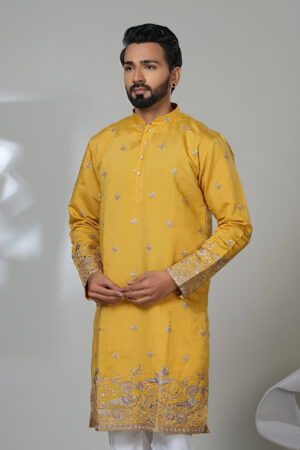 Man wearing a mustard yellow embroidered kurta with silver floral detailing, paired with white pants and black shoes, standing in a modern studio setup.