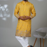 Man wearing a mustard yellow embroidered kurta with silver floral detailing, paired with white pants and black shoes, standing in a modern studio setup.