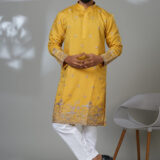 Man wearing a mustard yellow embroidered kurta with silver floral detailing, paired with white pants and black shoes, standing in a modern studio setup.