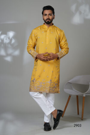 Man wearing a mustard yellow embroidered kurta with silver floral detailing, paired with white pants and black shoes, standing in a modern studio setup.
