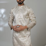 Man wearing a cream-colored embroidered kurta with subtle floral patterns, paired with white trousers and black shoes, standing in a studio setting.