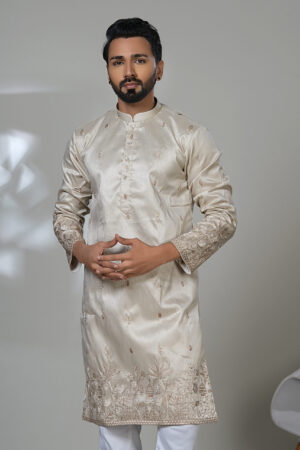Man wearing a cream-colored embroidered kurta with subtle floral patterns, paired with white trousers and black shoes, standing in a studio setting.