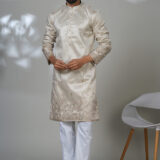 Man wearing a cream-colored embroidered kurta with subtle floral patterns, paired with white trousers and black shoes, standing in a studio setting.