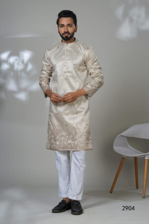 Man wearing a cream-colored embroidered kurta with subtle floral patterns, paired with white trousers and black shoes, standing in a studio setting.